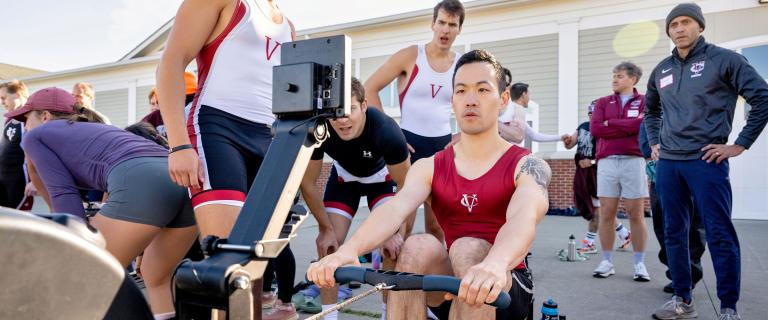 Several people stand around one person using a rowing machine.