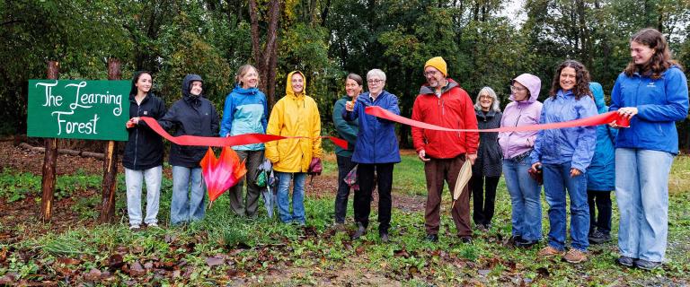 A group of people wearing rain jackets stand outdoors in front of a green sign that reads “The Learning Forest.” They smile as one person cuts a red ribbon during a rainy-day ribbon-cutting ceremony surrounded by trees.