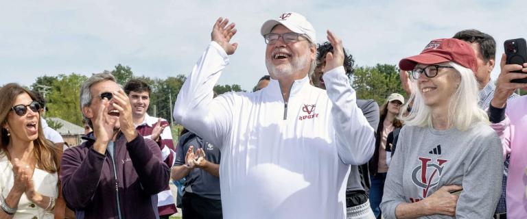 A crowd stands outdoors on grass, clapping and smiling. In the center, a person in a white quarter-zip pullover with a maroon “Rugby” logo raises both arms joyfully. People around them, some wearing sunglasses and Vassar-themed clothing, join in the celebration. The background includes trees and people partially visible in athletic uniforms.
