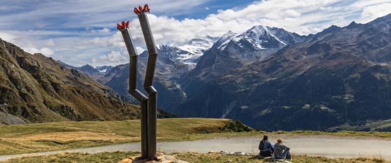 An abstract sculpture that looks like two crooked metal pipes stands outside next to a vast mountain range. Two people are sitting next to it.