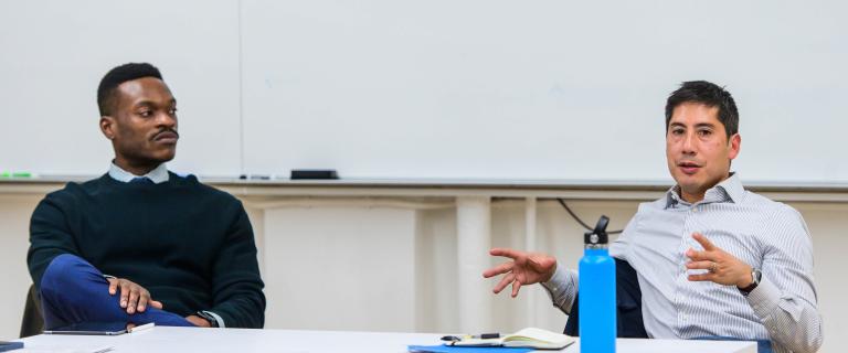  Two individuals seated at a table against a white background. The person on the left wears a dark sweater and blue pants, looking left. The person on the right wears a striped dress shirt, gesturing with both hands while speaking.