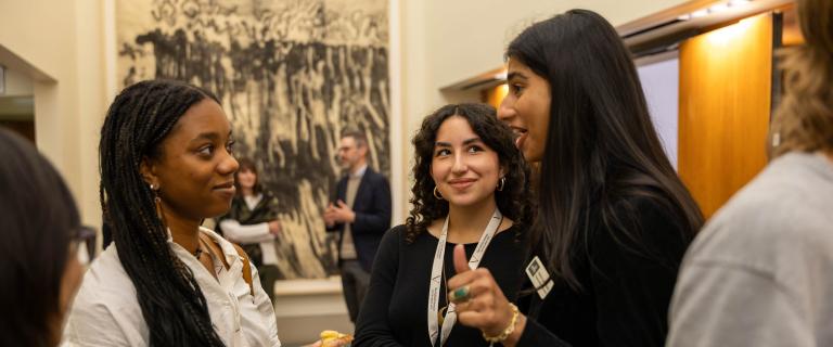  Three individuals in the foreground, engaged in a discussion at an indoor event. The person on the left has long braids and wears a white shirt. The person in the center smiles, wearing a black top and a lanyard. In the background, a large abstract black and white piece of art hangs on the wall, and other attendees are visible.