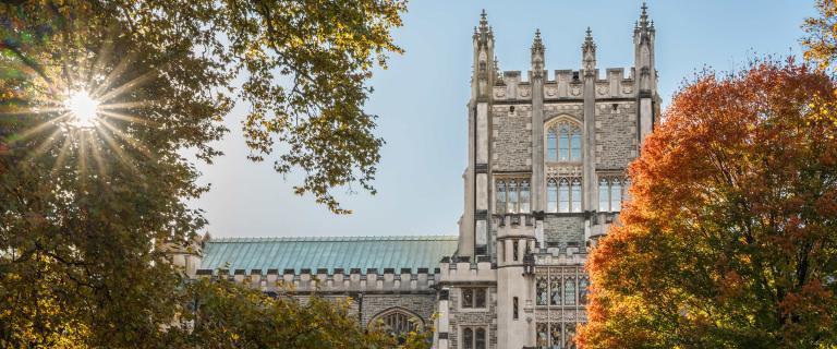 A historic, Gothic-style stone building with tall, ornate towers and large arched windows, partly framed by autumn trees with green and orange foliage. Sunlight shines through the leaves on the left, creating a bright starburst effect against a clear sky.