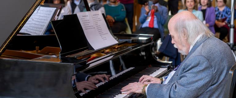 Person playing a grand piano, hands on the keys, with sheet music propped up and a blurred audience in the background.