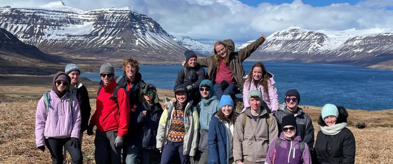 A large group of people bundled in jackets, hats, and gloves pose together on a grassy hillside with a stunning backdrop of snow-capped mountains and a deep blue fjord under a bright, partly cloudy sky.