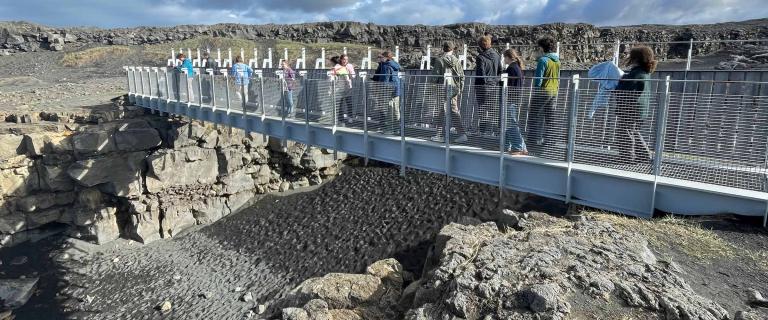 A group of people walk across a long metal footbridge that spans a rocky rift with dark sand below.