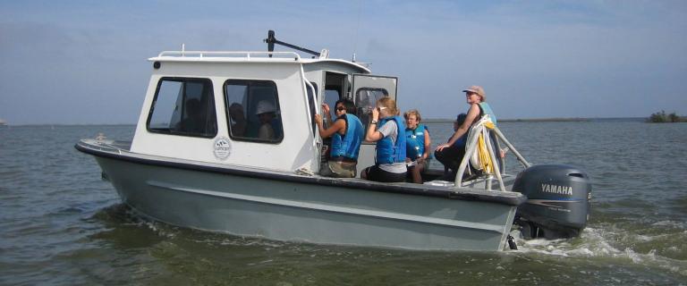 A small research boat moves across calm water, carrying several people wearing blue life vests.