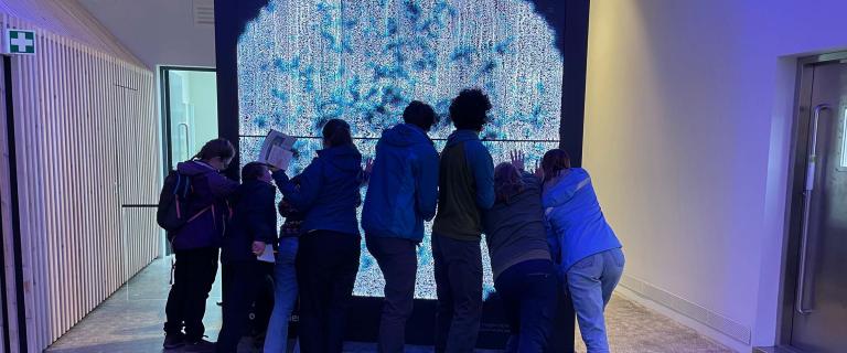 A group of people stand closely together in a dimly lit hallway, facing a large vertical screen glowing with bright blue, white, and teal abstract patterns.