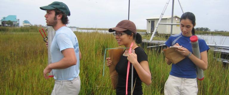 Three people stand in a grassy coastal marsh conducting fieldwork holding miscellaneous supplies with a dock and small building near the water in the background.