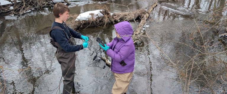 Two people stand in a shallow, cold stream wearing waders and gloves, handling cables and a handheld device as part of water-quality fieldwork. 