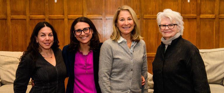 Four individuals standing side-by-side indoors, posing for a group portrait. The background features a plush sofa and paneled wooden walls. They are all dressed in business casual attire, smiling directly at the camera.