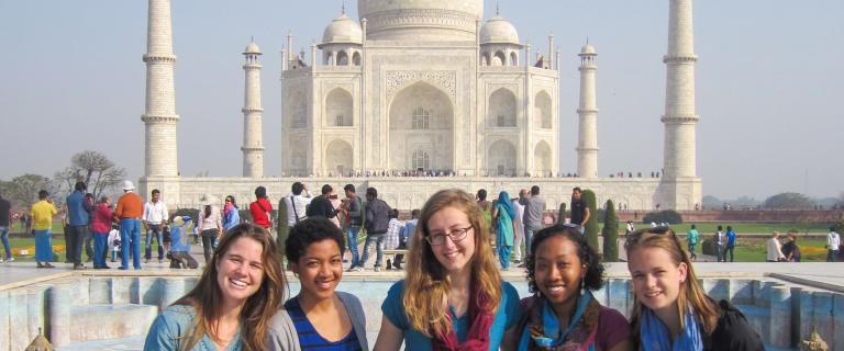 Four students smiling in the foreground with the Taj Mahal in the background, surrounded by other visitors on the grounds.