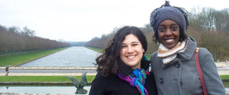 Two students smiling outdoors in winter coats and scarves, standing in front of a long reflecting canal lined with bare trees.
