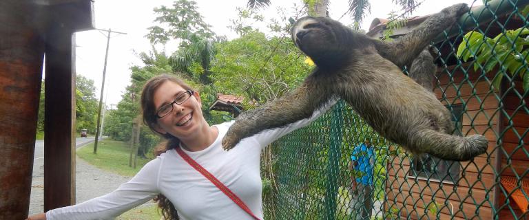 Smiling student posing beside a chain-link fence as a sloth stretches across the top of the fence with its limbs extended.