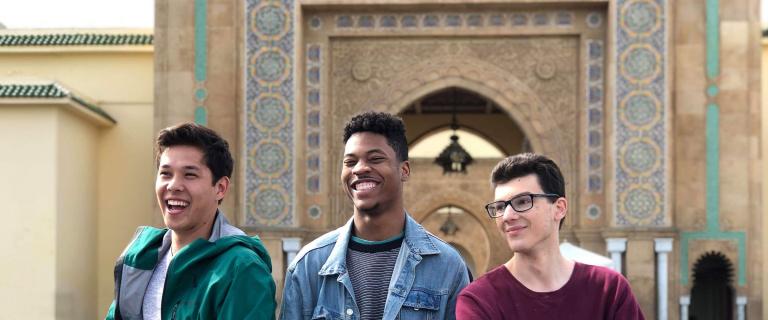 Three students smiling outdoors in front of an ornate tiled archway with Moroccan-style geometric patterns.