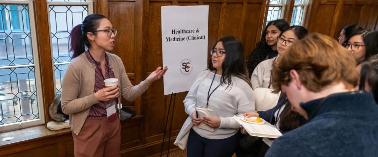 A speaker gestures while addressing a small group of students wearing name badges at a networking event near a “Healthcare & Medicine (Clinical)” sign.