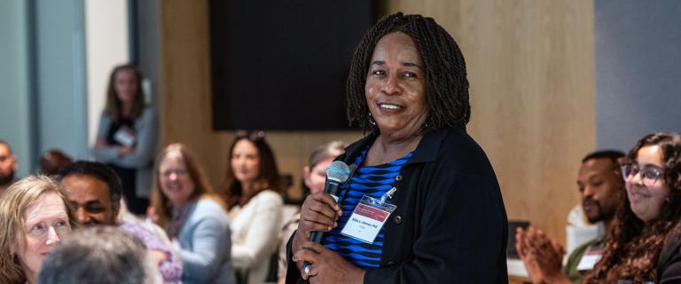 Person standing and speaking amid a seated smiling crowd.