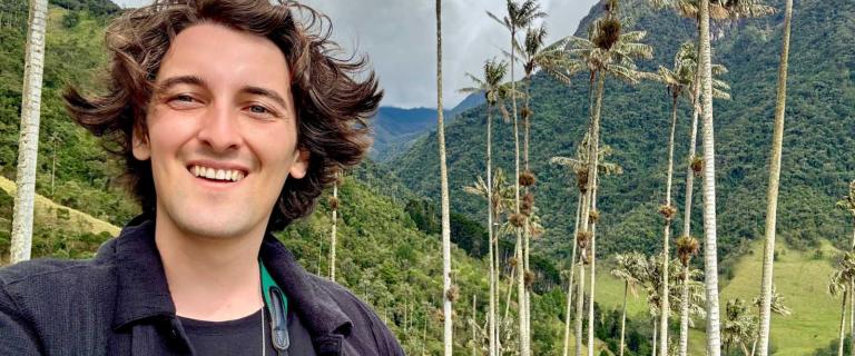 A person stands in a lush, mountainous landscape, surrounded by tall, slender trees. Bright green foliage contrasts with the cloudy sky, which features varying shades of gray. The person has brown hair, and they wear a dark, open jacket over a black shirt.