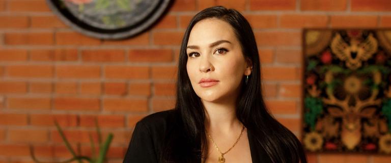 Headshot of Kali Fajardo-Anstine in front of a brick wall with artwork hanging on it.