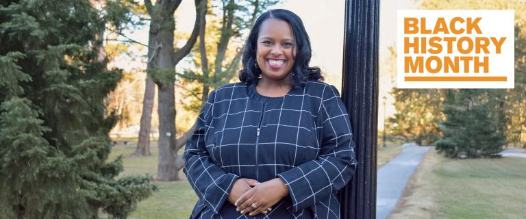 A smiling individual in a dark, grid-patterned jacket leans against a black post outdoors. Behind them is a park-like setting with trees and a walkway. The top right corner features a white graphic with the orange text "Black History Month."