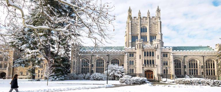Winter view of a Gothic-style stone campus building with a central tower and arched windows, surrounded by snow-covered trees and pathways, with a person walking across the snowy lawn in the foreground.