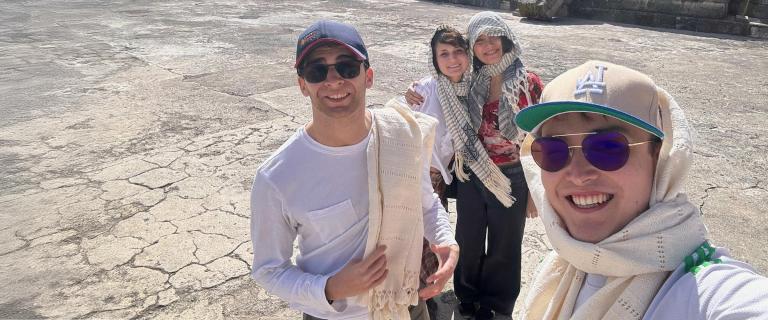Four people smiling for a selfie outdoors on a sunny day, wearing hats, sunglasses, and light scarves, standing on a wide stone surface that appears to be part of a historic site.
