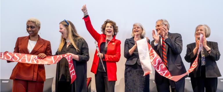 Group of smiling people hold a large ribbon with the word "Vassar" on it that has been cut in half.