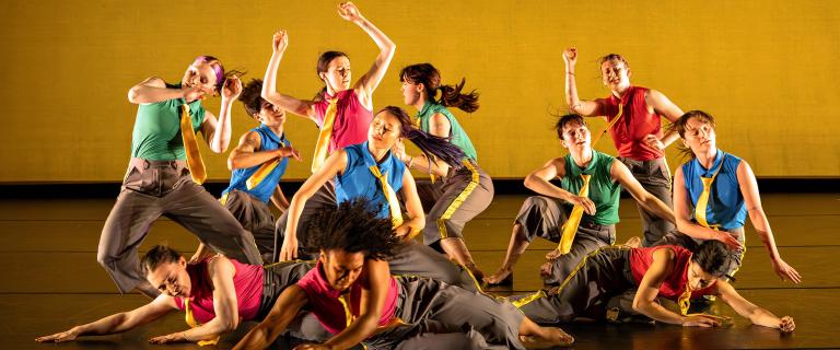 A group of dancers performs an energetic, synchronized routine on stage against a yellow backdrop, wearing colorful sleeveless tops, gray pants, and yellow ties.