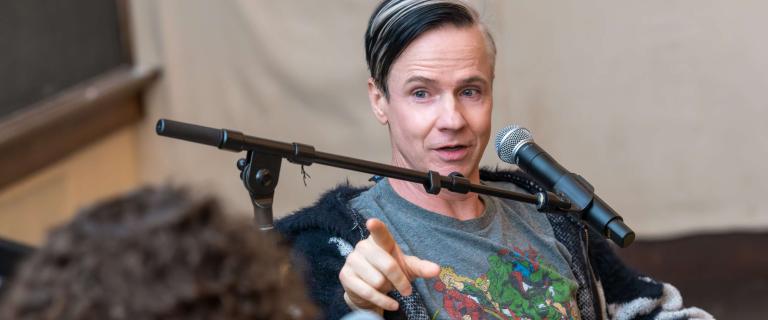 Actor/Director John Cameron Mitchell, a person with short dark hair, speaks in front of a microphone.