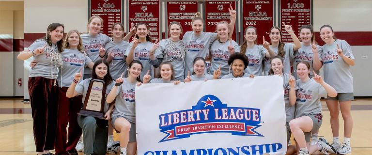 A group of people in athletic gear hold a large banner that says Liberty League Championship, with their index fingers raised.