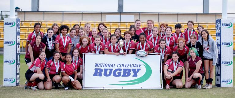 A group of people wearing athletic gear and medals around their necks pose in a goalpost with a large sign reading National Collegiate Rugby.