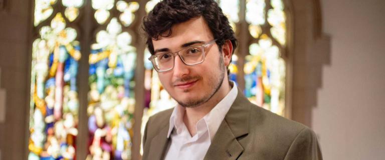 Bekir Hozdic ’26 stands in the Vassar library, with a stained glass window in the background. They are wearing glasses and a brown jacket.
