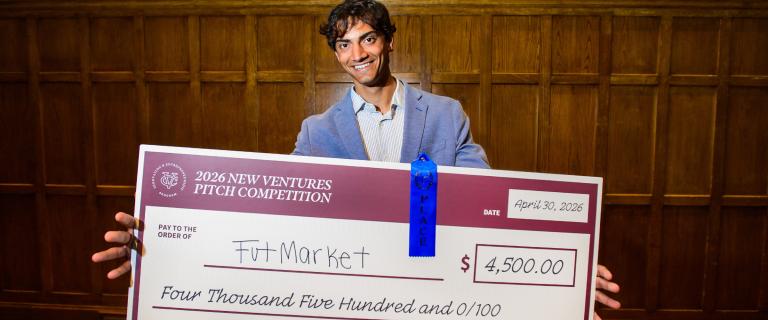 A smiling person holds a large first-place check for $4,500 from the 2026 New Ventures Pitch Competition while standing in front of a wood-paneled wall.
