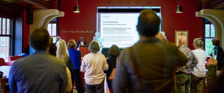 Audience members stand and listen to Dean of the Faculty, Demetrius Eudell, toasting at an event in the Class of 1951 reading room with a large presentation screen in the background.
