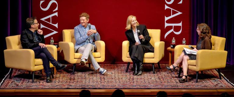 Four individuals sit in yellow armchairs on a stage during a panel discussion. Behind them is a red backdrop with the word VASSAR printed vertically. The stage is covered with a patterned rug, and some individuals hold microphones while talking.