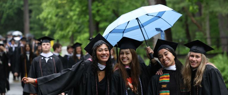 A smiling group of graduates in colorful Commencement attire and an umbrella.