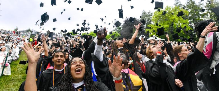 Graduates celebrate during commencement by tossing caps into the air while cheering and reaching upward, with a large crowd of attendees in rain ponchos gathered on the lawn in the background.