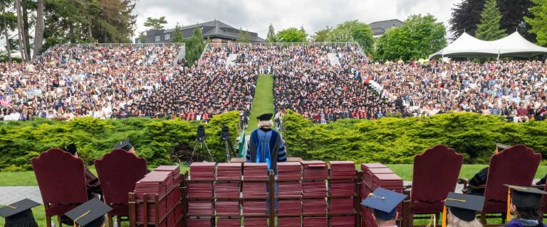 A commencement speaker in cap and gown speaking behind a lectern before a large outdoor audience.