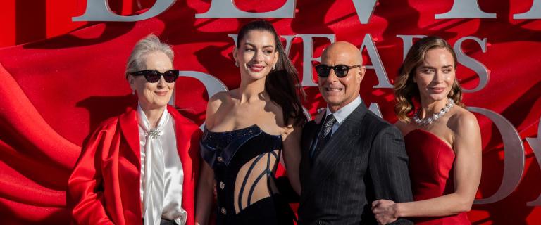 Four people posing together on a red carpet in formal attire in front of a bright red event backdrop with large lettering. Two people wear red outfits, one person wears a dark suit and sunglasses, and one person wears a dark evening gown. Strong sunlight creates dramatic shadows across the backdrop and clothing.