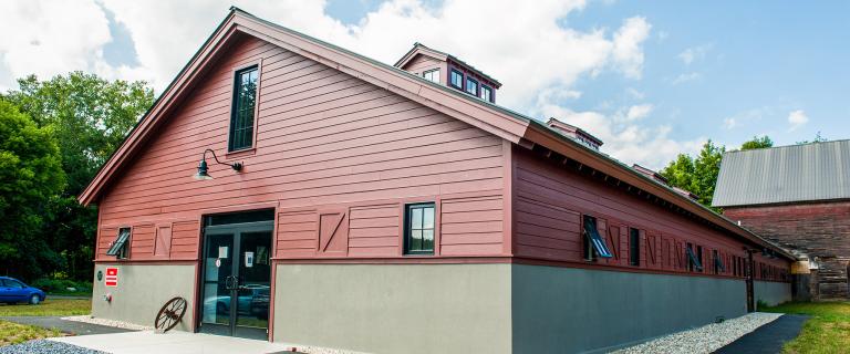 A large barn-style research and education facility with red siding and a gray foundation sits amid a natural landscape. The building features tall windows, dormers, and a main entrance beneath a partly cloudy sky.