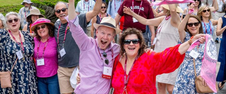 Participants in a campus reunion parade smile, wave, and celebrate while walking together outdoors. Several people wear Vassar reunion lanyards and name badges, with colorful clothing, hats, and scarves adding to the festive atmosphere. A crowd of alumni follows behind along a landscaped campus pathway.