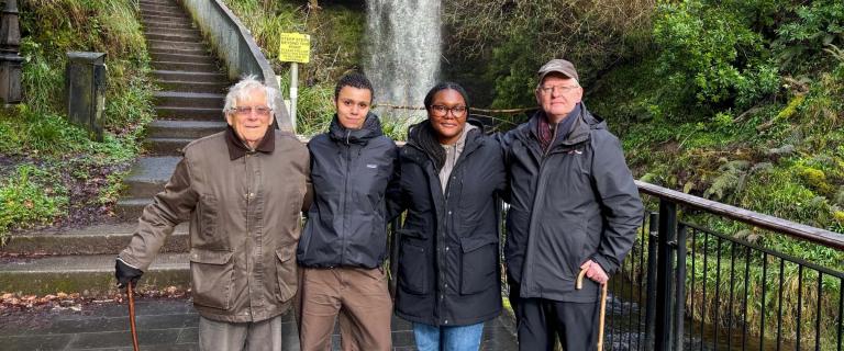 Four people stand outdoors, with foliage and a stone staircase behind them.