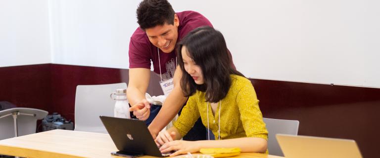 Two conference participants collaborate at a table, with one person pointing at a laptop screen while the other works on the computer. Both wear conference name badges, and reusable water bottles and laptops are visible in the bright classroom setting, suggesting a hands-on workshop or collaborative learning session.