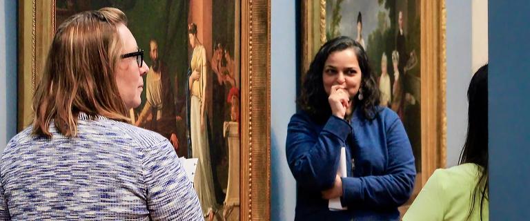Two people converse in a gallery with framed paintings displayed behind them.