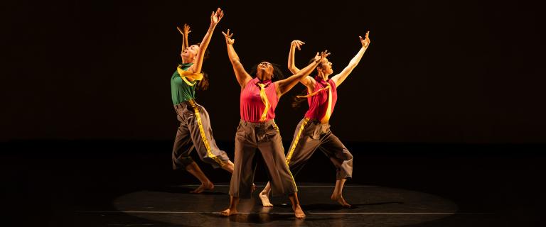 Three dancers perform on a dark stage under warm spotlights, striking expressive poses with raised arms. They wear brightly colored sleeveless tops with flowing yellow scarves and cropped gray pants accented with yellow stripes, creating a dynamic, synchronized composition.