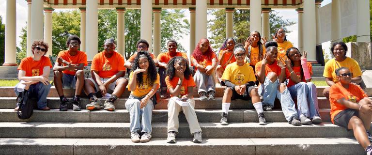 A group of students wearing orange and yellow shirts sits together on outdoor steps.