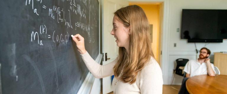 A person writes complex equations on a blackboard while another person sits at a table in the background watching.