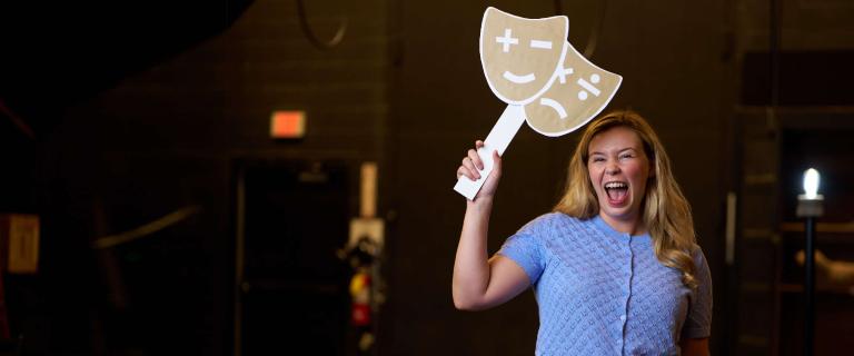 A figure holds two large, playful masks on sticks, one featuring a smiley face and the other a neutral face, raised high in an energetic gesture. The figure is dressed in a light blue, short-sleeved top with a textured pattern and appears to be laughing. The background features dark walls with faint outlines of stage equipment and dim lighting that enhances the focused spotlight illuminating the figure. The overall composition conveys a lively and interactive atmosphere.