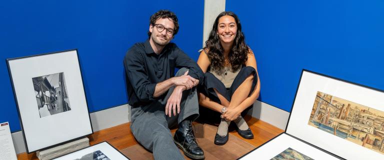 Two people sit smiling on the floor of a gallery surrounded by framed artwork in front of a bright blue wall.