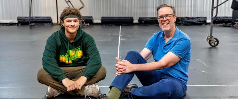 Two people sit cross-legged and smile on a black stage floor inside a theater space with scaffolding in the background.